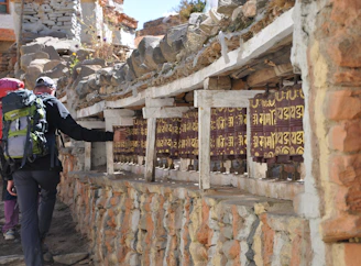 A person with a backpack is spinning prayer wheels set into a stone wall in a rustic outdoor setting. The prayer wheels feature inscriptions in a script format and are lined up under a shelf covered with stones. The scene exudes a sense of tranquility and tradition, with sunlight illuminating the area.