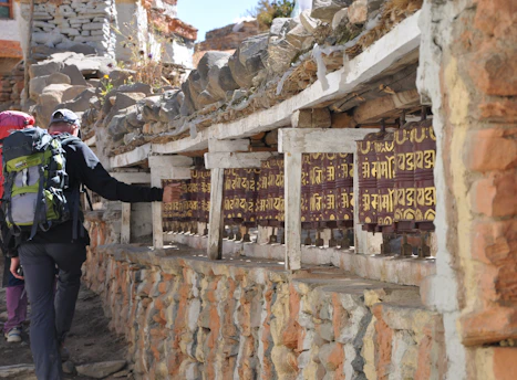 A person with a backpack is spinning prayer wheels set into a stone wall in a rustic outdoor setting. The prayer wheels feature inscriptions in a script format and are lined up under a shelf covered with stones. The scene exudes a sense of tranquility and tradition, with sunlight illuminating the area.
