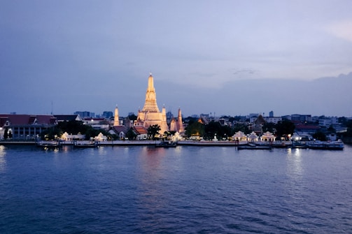A beautifully illuminated temple stands out against a twilight sky, reflecting its golden light onto the still waters of a river. Traditional Thai architecture with ornate designs is evident, as the temple is surrounded by smaller buildings and trees in the cityscape.
