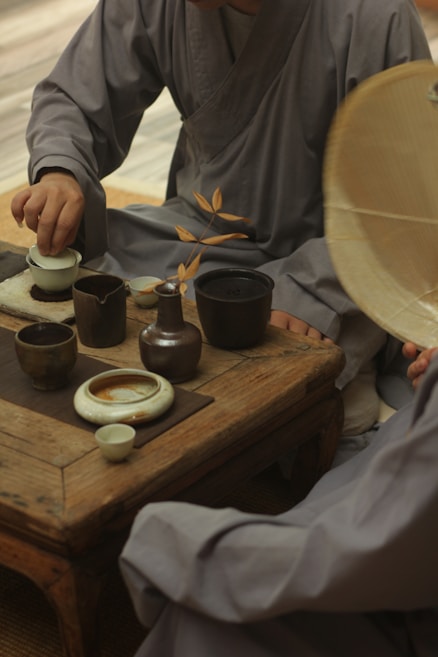 Two people dressed in traditional attire are seated around a wooden table with traditional tea utensils including a teapot, cups, and a branch of dried leaves. One person is pouring tea while another holds a round, wicker object, possibly a hat.