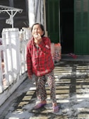 An elderly person stands outside in the sunlight, wearing a red plaid shirt and patterned pants. They are on a concrete surface, possibly a balcony or patio, with a white railing. In the background, there are dark green shutters and a partially open door leading indoors. Some household items, such as a drying rack with hangers and plastic bags, are visible.