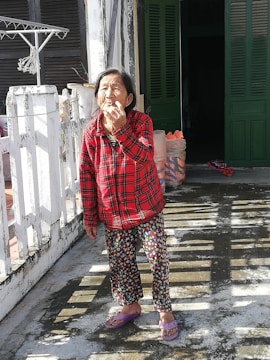 An elderly person stands outside in the sunlight, wearing a red plaid shirt and patterned pants. They are on a concrete surface, possibly a balcony or patio, with a white railing. In the background, there are dark green shutters and a partially open door leading indoors. Some household items, such as a drying rack with hangers and plastic bags, are visible.