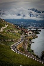 A scenic landscape of a winding road and railway running parallel along a hillside covered in lush vineyards. Quaint houses and buildings are nestled along the slopes with a lake on the right. Snow-capped mountains in the background are partially obscured by low-hanging clouds, creating a dramatic and picturesque setting.