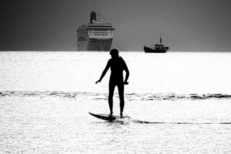 A silhouetted person is paddleboarding on calm water, with a large cruise ship and a smaller boat in the distance. The scene is set during a low-light time, possibly dawn or dusk, creating a serene atmosphere.