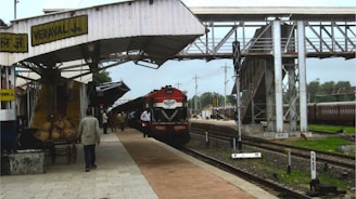 A train station platform with a red and black locomotive parked at the station. The platform is partially covered by a metal canopy with a sign reading 'VERAVAL JN'. Several people are walking along the platform, and there are items loaded on a cart. A pedestrian overpass is visible, and there are green trees in the background.