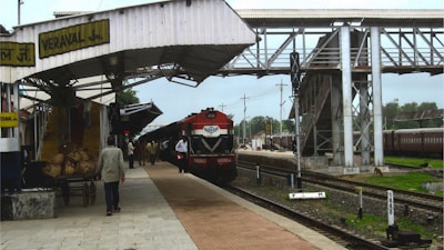 A train station platform with a red and black locomotive parked at the station. The platform is partially covered by a metal canopy with a sign reading 'VERAVAL JN'. Several people are walking along the platform, and there are items loaded on a cart. A pedestrian overpass is visible, and there are green trees in the background.