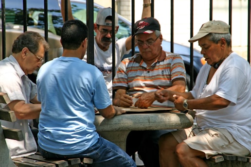 A group of five men are sitting around a small circular stone table, engaged in a game that involves cards or tiles. They are outdoors, possibly in a park, as metal railings and trees are visible in the background. The men are of various ages, wearing casual clothing like t-shirts and caps, and appear focused on their game.