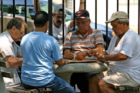 A group of five men are sitting around a small circular stone table, engaged in a game that involves cards or tiles. They are outdoors, possibly in a park, as metal railings and trees are visible in the background. The men are of various ages, wearing casual clothing like t-shirts and caps, and appear focused on their game.