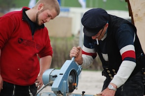 Two men are focused on using a blue power tool, likely a saw. One man in a red sweatshirt watches attentively, while the other in a cap and navy sweater operates the machine. Their expressions suggest concentration. The background is blurred, indicating an outdoor setting possibly related to construction or woodworking.