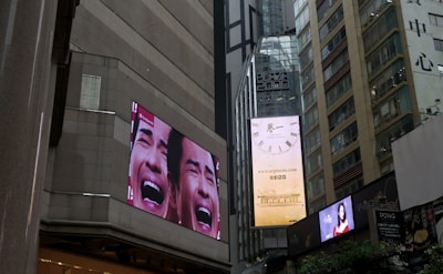 Tall urban buildings with large digital billboards displaying advertisements. One billboard shows a close-up of a face with an expressive emotion, and another displays a clock advertisement.