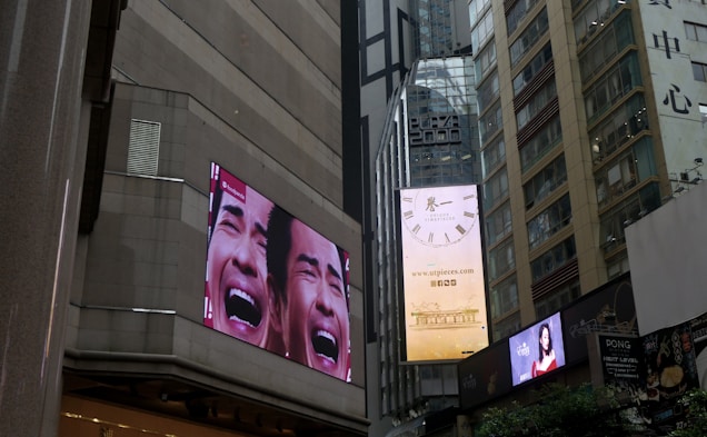 Tall urban buildings with large digital billboards displaying advertisements. One billboard shows a close-up of a face with an expressive emotion, and another displays a clock advertisement.