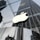 A large, illuminated apple logo suspended in front of a modern glass building. The glass walls reflect the surrounding architecture, including a tall skyscraper and other urban structures, creating a sleek and modern cityscape.