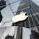 A large, illuminated apple logo suspended in front of a modern glass building. The glass walls reflect the surrounding architecture, including a tall skyscraper and other urban structures, creating a sleek and modern cityscape.
