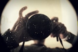 A close-up view of a person holding a camera lens, with the silhouette and focus on the lens, creating a mysterious and introspective atmosphere. The image is slightly obscured and blurry, enhancing the sense of depth and texture.