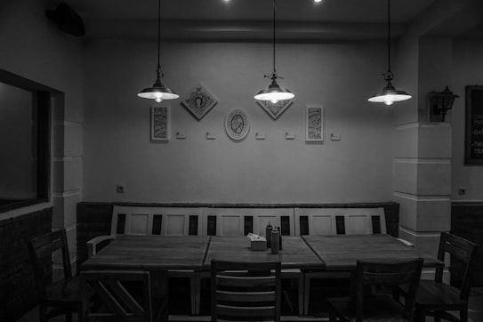 A monochrome interior of a dining area with a wooden table and several chairs. Three hanging lights illuminate the space, casting shadows on the walls. Decorative frames and wall art are arranged symmetrically above the bench seating along the back wall.