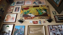 A collage of vintage and retro-style posters and advertisements covers a wall. Various framed artworks depict classic brand logos, drinks, and pin-up art. Notable elements include a large painting featuring bottles and a lime, a retro advertisement with a glamorous woman, and a collection of smaller, nostalgic images.