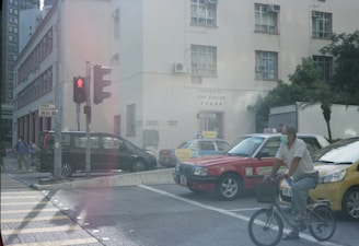 A busy urban street scene with a traffic light at a crossroads. A bicyclist wearing a mask is riding past two taxis parked near a building marked 'Hong Kong Fire Station'. Several pedestrians in masks are visible in the background near a black van. The atmosphere is bright with some greenery in the area.
