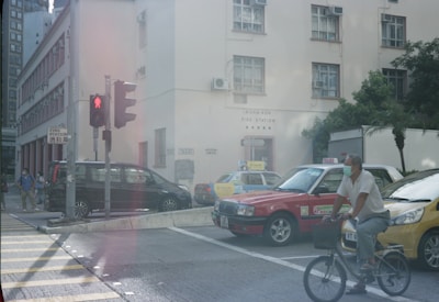 A busy urban street scene with a traffic light at a crossroads. A bicyclist wearing a mask is riding past two taxis parked near a building marked 'Hong Kong Fire Station'. Several pedestrians in masks are visible in the background near a black van. The atmosphere is bright with some greenery in the area.