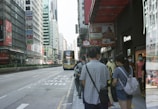 A busy urban street scene with a group of people standing on a sidewalk. Many of them are wearing face masks, suggesting awareness of health concerns. A double-decker bus is approaching, and tall buildings line the street, which is adorned with various signs and advertisements. The atmosphere is typical of a bustling city environment.
