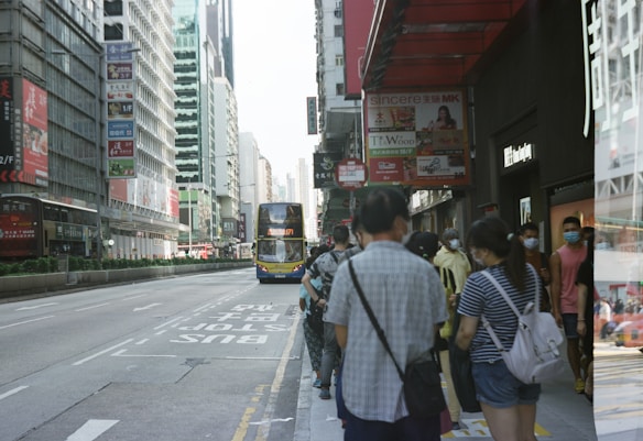 A busy urban street scene with a group of people standing on a sidewalk. Many of them are wearing face masks, suggesting awareness of health concerns. A double-decker bus is approaching, and tall buildings line the street, which is adorned with various signs and advertisements. The atmosphere is typical of a bustling city environment.