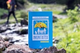 A book titled 'Imunitatea' is positioned in the foreground, with a vibrant blue cover featuring an illustration of a family under a yellow umbrella. The background shows a blurred natural setting with a hiker walking, highlighting greenery and a small stream.