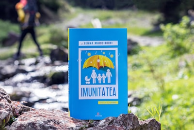 A book titled 'Imunitatea' is positioned in the foreground, with a vibrant blue cover featuring an illustration of a family under a yellow umbrella. The background shows a blurred natural setting with a hiker walking, highlighting greenery and a small stream.