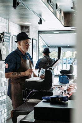 A man wearing a brown leather apron and a hat stands at a counter, likely in a butcher shop. He appears focused, possibly preparing or weighing meat. Another person in similar attire can be seen in the background, also working at a counter. The shop interior has a modern design with white subway tiles and natural light streaming in from the large window.