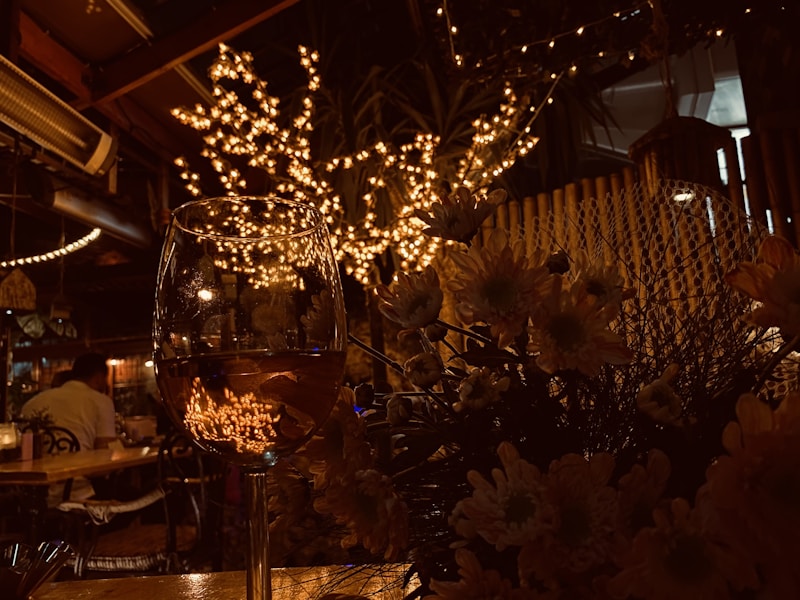 A warmly lit, intimate restaurant setting with string lights illuminating the background. A wine glass in the foreground reflects the ambient lighting. Flowers add a decorative touch to the lower half of the image, while a person is seated at a table in the distance.