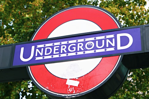 A sign featuring a bold red circle with a blue rectangle intersecting it, displaying the word 'Underground' in white letters. The background consists of green foliage, likely from trees.