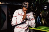 A person with braided hair wearing a white long-sleeve shirt with red text patterns is holding their hands together in a praying gesture. The background features colorful, abstract graffiti art.