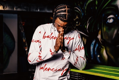 A person with braided hair wearing a white long-sleeve shirt with red text patterns is holding their hands together in a praying gesture. The background features colorful, abstract graffiti art.