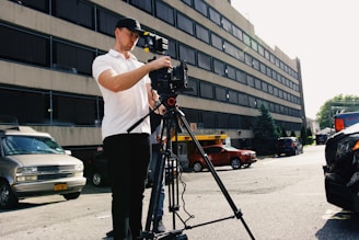 A man wearing a white shirt and black cap is operating a professional camera mounted on a tripod in an urban setting. The background features a large building with multiple floors and windows. Several vehicles are parked along the street, including a van and a red car. The lighting suggests a sunny day.