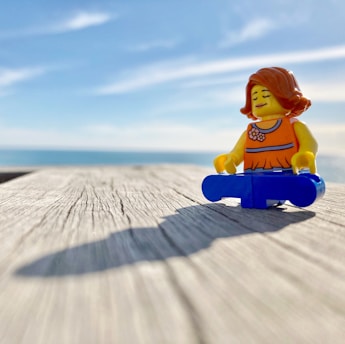 A LEGO figurine is sitting in a meditative pose on a wooden surface with a vast ocean and clear blue sky in the background. The figurine has orange hair, a yellow face, and is wearing an orange top and blue legs.