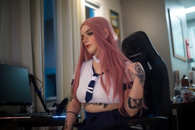 A person with long pink hair is sitting on a gaming chair, wearing a sailor-style outfit with tattoos visible on their arms. The room has dim lighting and there is a desk with a computer monitor and other tech equipment nearby. The interior has neutral-toned walls and decorations.