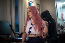 A person with long pink hair is sitting on a gaming chair, wearing a sailor-style outfit with tattoos visible on their arms. The room has dim lighting and there is a desk with a computer monitor and other tech equipment nearby. The interior has neutral-toned walls and decorations.