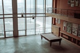 A large room with tall windows allowing plenty of natural light. The room features a pool table in the center and several posters on the back wall. The floor appears to be tiled, and there are a few decorative hanging lanterns. A railing on the upper floor overlooks the room.