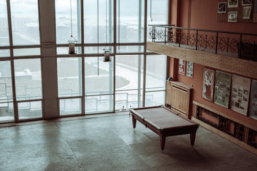 A large room with tall windows allowing plenty of natural light. The room features a pool table in the center and several posters on the back wall. The floor appears to be tiled, and there are a few decorative hanging lanterns. A railing on the upper floor overlooks the room.
