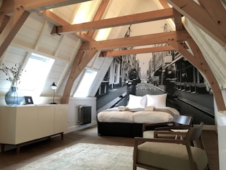A cozy attic bedroom with a slanted wooden beam ceiling featuring an artistic black and white mural of a street scene as the backdrop. The room includes a comfortable bed with white linens, a modern lamp, a vase with branches on a cabinet, a radiator, and minimalistic furniture. Natural light pours in from ceiling windows, creating a serene and inviting atmosphere.