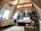 A cozy attic bedroom with a slanted wooden beam ceiling featuring an artistic black and white mural of a street scene as the backdrop. The room includes a comfortable bed with white linens, a modern lamp, a vase with branches on a cabinet, a radiator, and minimalistic furniture. Natural light pours in from ceiling windows, creating a serene and inviting atmosphere.