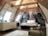 A cozy attic bedroom with a slanted wooden beam ceiling featuring an artistic black and white mural of a street scene as the backdrop. The room includes a comfortable bed with white linens, a modern lamp, a vase with branches on a cabinet, a radiator, and minimalistic furniture. Natural light pours in from ceiling windows, creating a serene and inviting atmosphere.