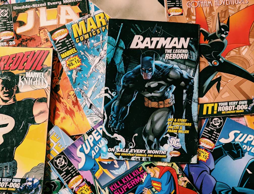 A collection of various comic books scattered on a surface. The comic books feature popular superheroes like Batman and Superman. The covers display colorful, dynamic illustrations with bold titles and iconic characters in action poses.