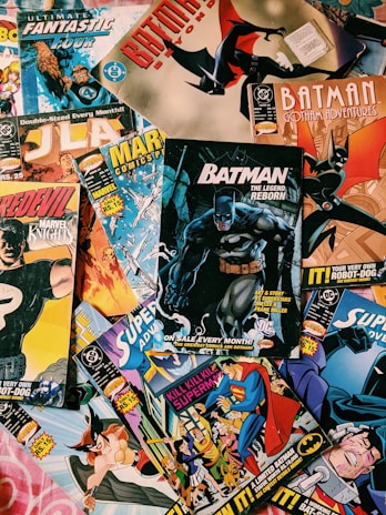 A collection of various comic books scattered on a surface. The comic books feature popular superheroes like Batman and Superman. The covers display colorful, dynamic illustrations with bold titles and iconic characters in action poses.
