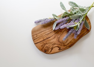 A wooden cutting board with a grain pattern lies on a light-colored surface, adorned with sprigs of lavender featuring green leaves and purple flowers.