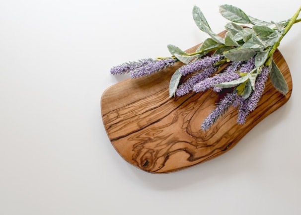 A wooden cutting board with a grain pattern lies on a light-colored surface, adorned with sprigs of lavender featuring green leaves and purple flowers.