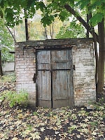 An old wooden door with peeling paint is set within a brick structure, surrounded by trees and dense foliage. Fallen leaves cover the ground, creating a rustic and natural appearance. The bricks appear weathered, suggesting age and exposure to the elements.