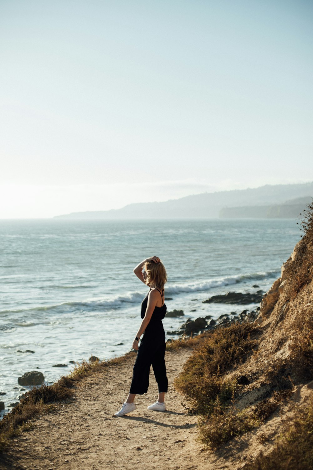 woman in black long sleeve dress standing on brown rock near body of water during daytime