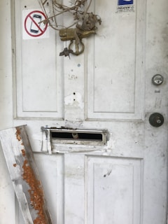A worn and weathered white door with visible damage, including a broken mail slot and worn-out lock. A piece of wood with rust-colored debris leans against the door. A dried-up wreath hangs from the top, and a sign prohibiting mobile phones is displayed near the top left corner.
