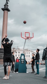 A group of people gathered in a public square, playing basketball. One individual is in the process of shooting the ball toward the hoop, which is set up with a backdrop of a large column and classical architecture. The sky is overcast, contributing to a lively yet relaxed atmosphere.