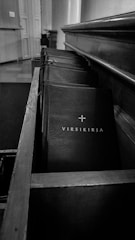 A row of hymn books is neatly arranged in a wooden pew or shelf inside a church or chapel. The lighting is subdued, creating a serene and solemn atmosphere. The main focus is on a book with the title 'Virsikirja' in Finnish, which translates to 'Hymn Book' in English.