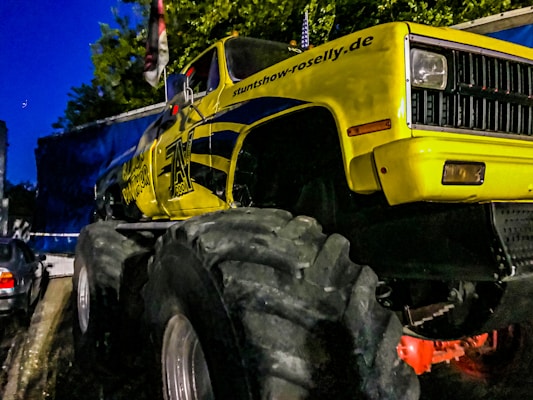 A large, yellow monster truck with oversized tires is parked on a street. The truck has various decals and text on it, including a website address. There is another vehicle to the left and a blue structure in the background along with some greenery. It appears to be evening as the sky is dark.